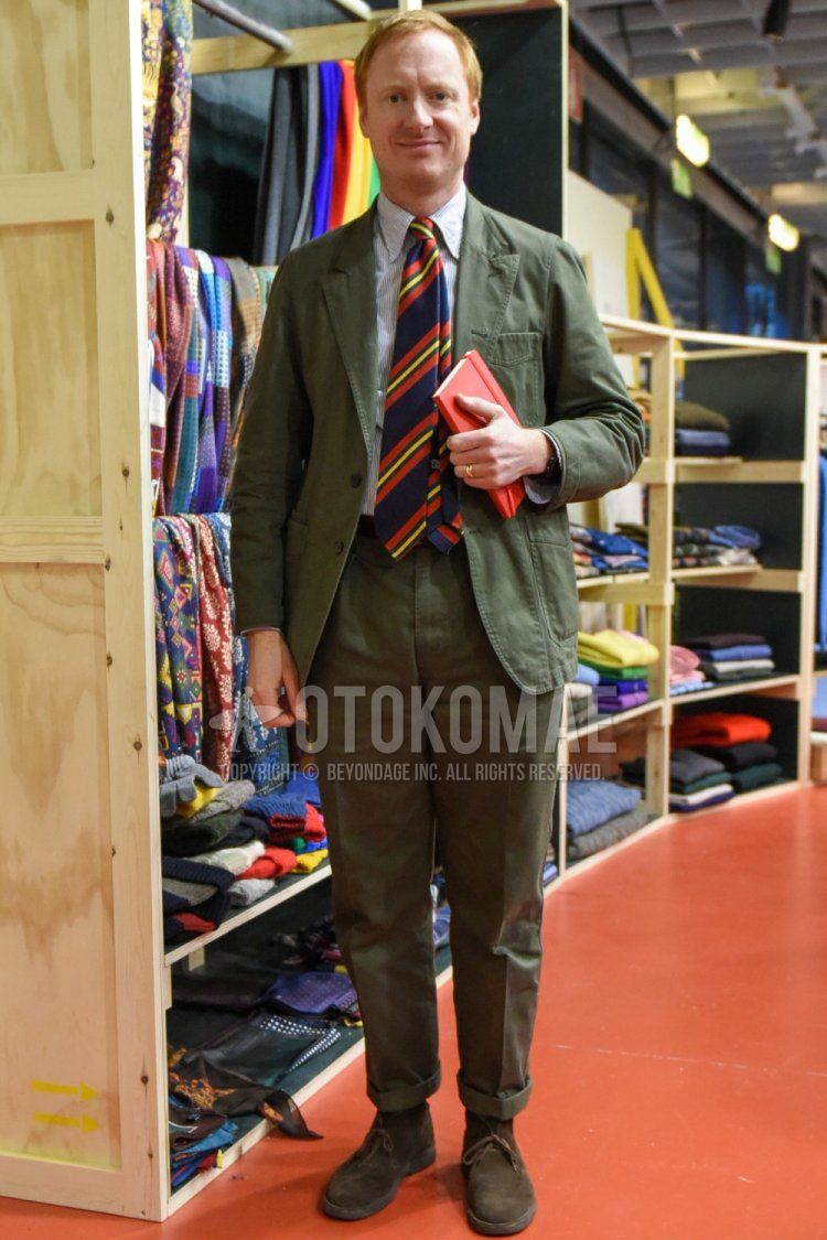 Men's spring and fall coordinate and outfit with light blue striped shirt, suede brown chukka boots, olive green solid color suit, and multi-colored regimental tie.