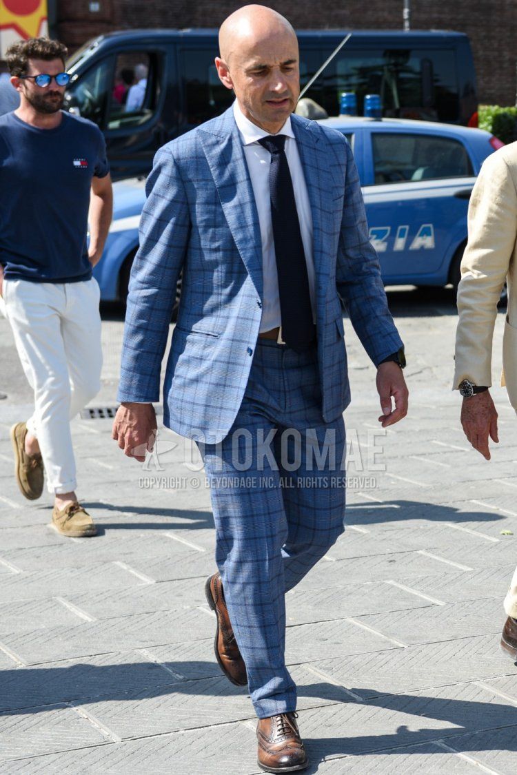 Men's spring and fall coordinate and outfit with plain white shirt, plain brown leather belt, brown brogue shoes leather shoes, blue checked suit, and plain navy tie.