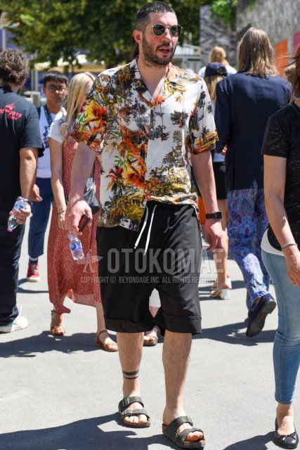 Summer men's coordinate and outfit with gold-black solid sunglasses, multi-colored botanical shirt, solid black shorts, and black leather sandals.
