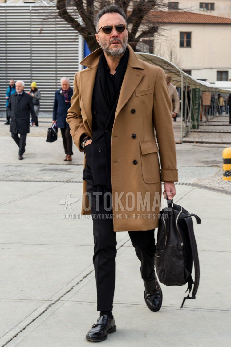 Men's fall/winter outfit with brown tortoiseshell sunglasses, solid black scarf/stall, solid beige Ulster coat, solid black socks, black monk shoe leather shoes, solid black backpack, solid black suit.