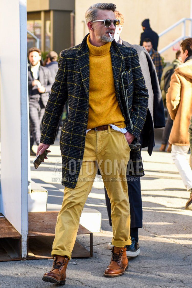 Men's fall/winter outfit with clear solid sunglasses, gray check chester coat, yellow solid turtleneck knit, brown solid leather belt, beige solid chinos, and brown work boots.