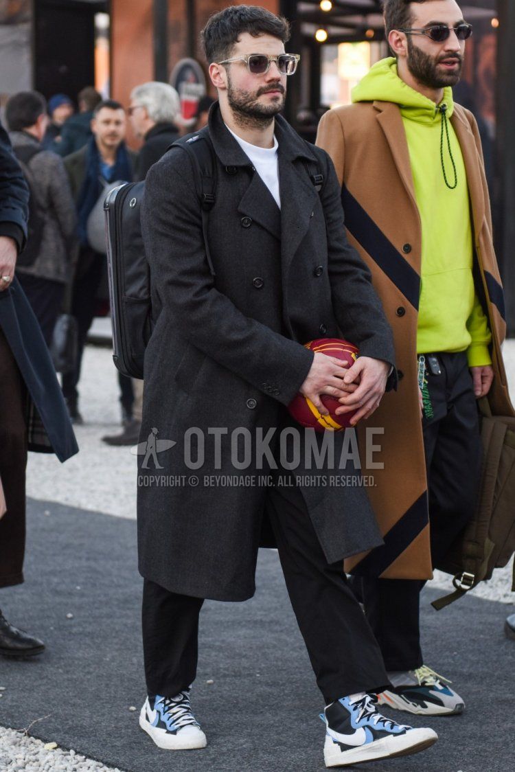 Men's fall/winter outfit and outfit with plain clear sunglasses, plain gray trench coat, plain white sweatshirt, plain gray slacks, plain gray cropped pants, Nike Sakai Blazer black, light blue and white high-cut sneakers, and plain black backpack.