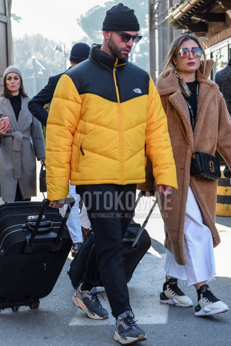 Men's winter/autumn coordinate and outfit with plain black knit cap, plain black sunglasses, The North Face yellow/black plain down jacket, plain black slacks, Adidas Easy Boost gray low-cut sneakers, and a plain black suitcase.