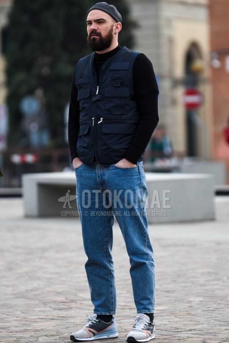Men's spring/fall outfit with plain gray cap, plain navy casual vest, solid black sweater, solid blue denim/jeans, solid black socks, and gray low-cut sneakers.