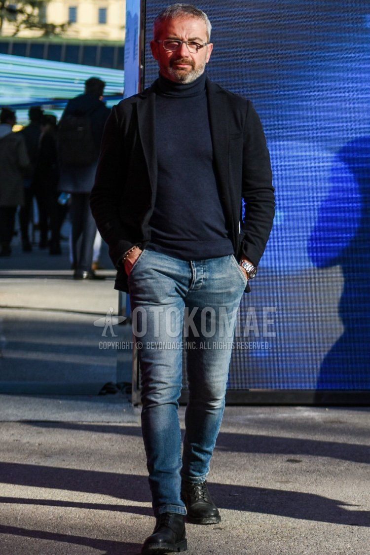 Men's spring/autumn coordination and outfit with plain silver glasses, plain black tailored jacket, dark gray plain turtleneck knit, solid blue denim/jeans, and black boots.
