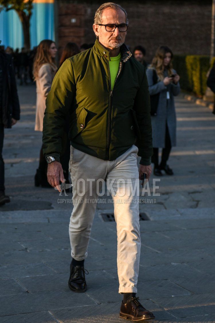 Men's fall/winter outfit with solid black glasses, solid olive green outerwear, solid yellow sweater, solid beige chinos, solid black socks, and brown plain toe leather shoes.
