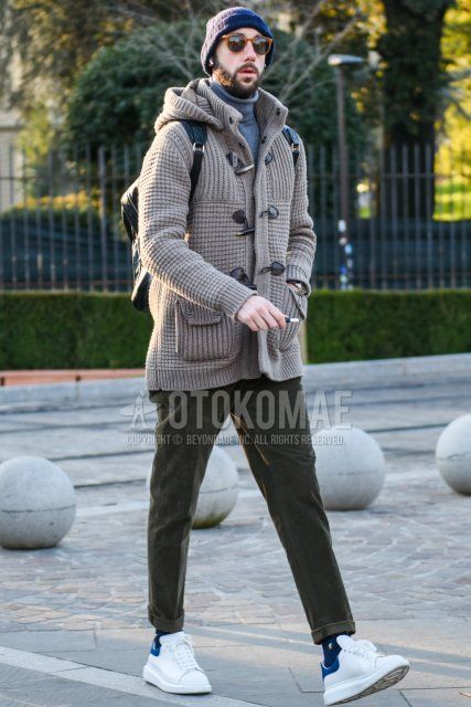 Men's fall/winter outfit with solid navy knit cap, solid brown sunglasses, solid beige duffle coat, solid gray turtleneck knit, solid olive green chinos, navy socks, white low-cut sneakers, and solid black backpack Outfit.