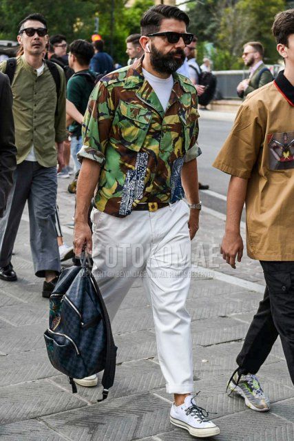 Men's spring/summer/fall outfit with plain black sunglasses, green/brown camouflage shirt jacket, plain white t-shirt, plain brown leather belt, plain white chinos, white low-cut sneakers, Louis Vuitton Damier gray/black bag backpack.