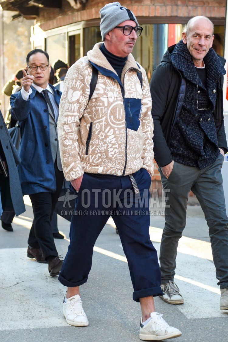 Men's fall/winter outfit with plain gray knit cap, plain black glasses, beige graphic fleece jacket, plain black turtleneck knit, plain navy chinos, and white low-cut sneakers by Golden Goose.