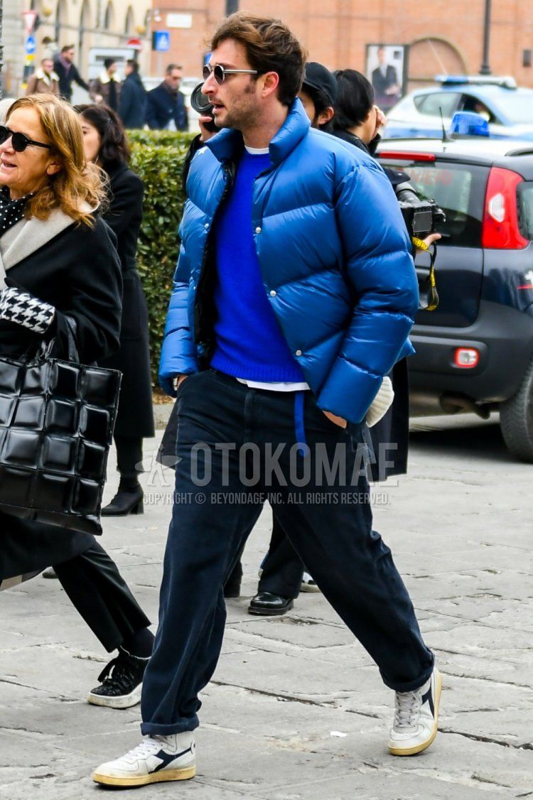 Winter men's coordinate and outfit with solid color sunglasses, solid color blue down jacket, solid color blue sweater, solid color navy chinos, solid color navy cotton pants, and white low-cut sneakers by Diadora.