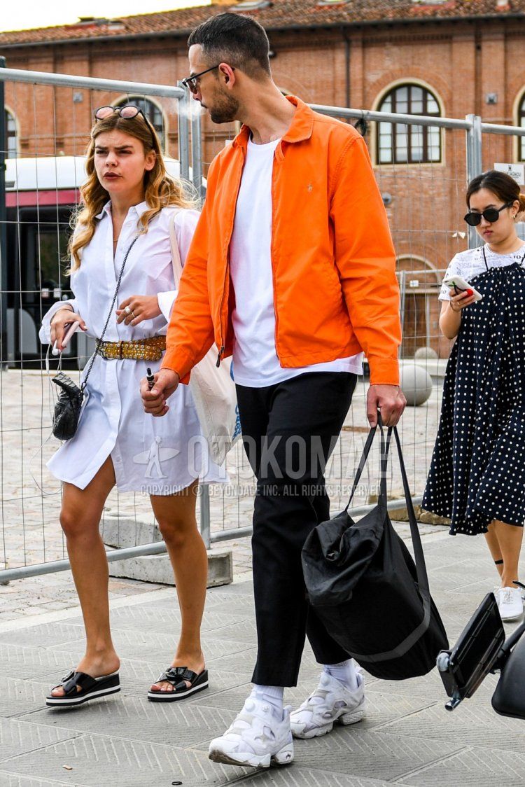 Men's spring/summer/fall outfit with solid color sunglasses, solid color orange swing top, solid color white t-shirt, solid color black ankle pants, solid color white socks, white low-cut Reebok Pump Hewley sneakers, and solid color black backpack.
