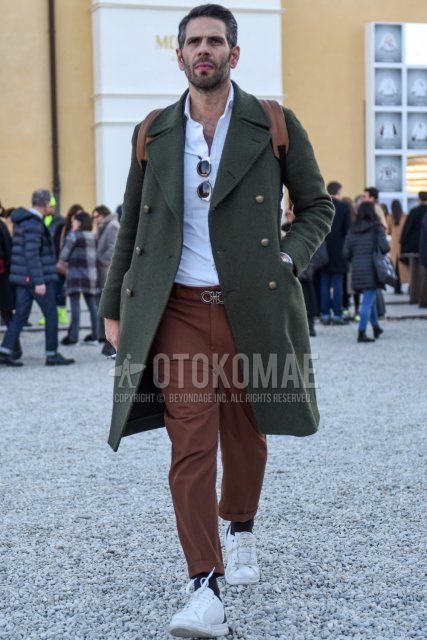 Men's fall/winter outfit with olive green solid color Ulster coat, white solid color shirt, brown solid color leather belt by Salvatore Ferragamo, brown solid color chinos, brown solid color ankle pants, black solid color socks, white low cut sneakers.