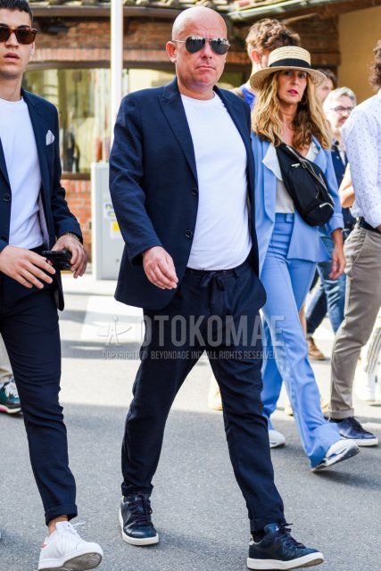 Men's spring and summer coordinate and outfit with teardrop solid beige sunglasses, solid white t-shirt, diadora navy low-cut sneakers, and solid navy suit.