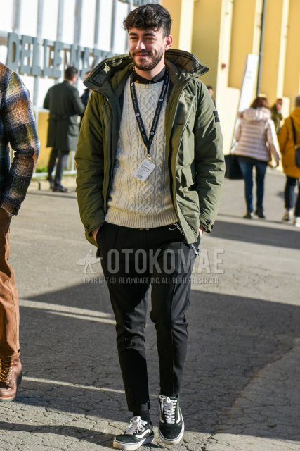 Men's fall/winter outfit with olive green solid color down jacket, solid color white sweater, solid color black shirt, solid color black slacks, solid color black ankle pants, solid color black socks, and Vans Old School black low cut sneakers.