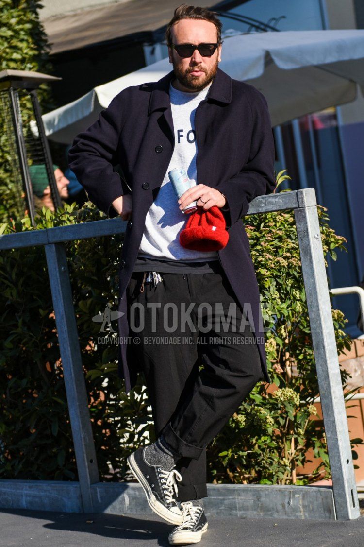 Men's fall/winter outfit with plain black sunglasses, plain navy stainless steel collar coat, gray graphic sweatshirt, plain black slacks, plain gray socks, and Converse Chuck Taylor black low-cut sneakers.