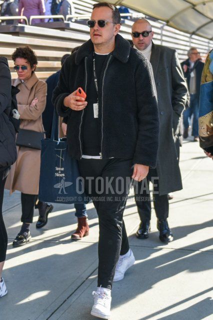 Clear plain sunglasses, plain black leather jacket (not riders), plain black sweater, plain black sweater, plain white t-shirt, plain gray slacks, plain gray ankle pants, plain white socks, Reebok Club Sea white low cut sneakers, gray graphic Tote bag with a men's fall/winter outfit.