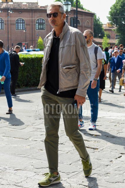 Men's spring and fall outfit with solid black sunglasses, solid beige outerwear, solid black polo shirt, solid olive green slacks, and Diadora olive green low-cut sneakers.