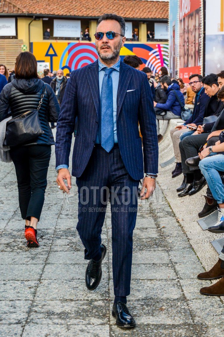 Men's spring/fall outfit with solid color sunglasses, solid color blue denim/chambray shirt, solid color black socks, black plain toe leather shoes, navy striped suit, and solid color blue tie.