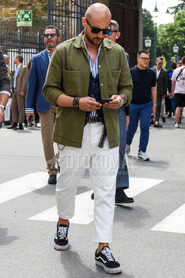 Spring and fall men's coordinate and outfit with Wellington plain black sunglasses, olive green plain shirt jacket, light blue and white striped shirt, navy plain gilet, black dotted leather belt, plain white ankle pants, and black low-cut Vans sneakers.