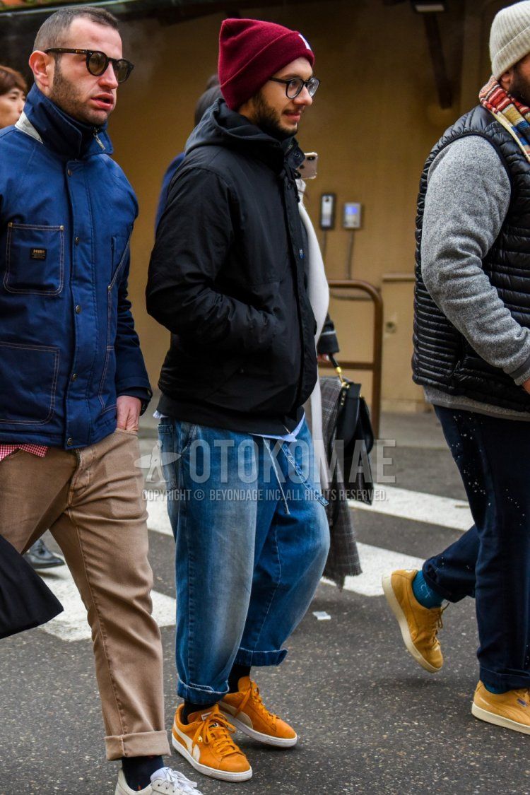 Men's spring/fall/winter coordinate and outfit with red solid knit cap, solid glasses, solid black windbreaker, solid blue denim/jeans, solid black socks, and Puma yellow low-cut sneakers.