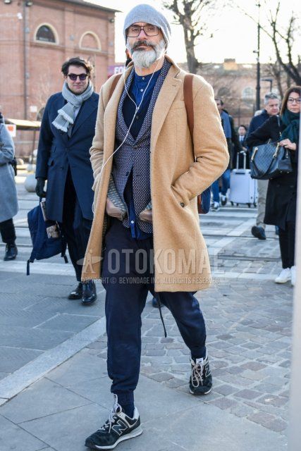 Solid gray knit cap, solid black glasses, solid beige chester coat, tailored jacket with gray top/inner, solid blue shirt with band collar, solid navy jogger pants/ribbed pants, solid navy socks, and New Balance gray low cut Men's fall/winter coordinate/outfit with sneakers.