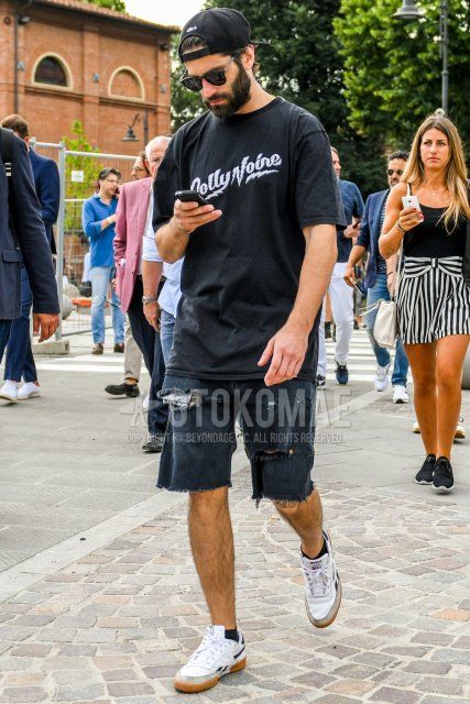 Summer men's coordinate and outfit with plain black baseball cap, plain black sunglasses, black graphic t-shirt, dark gray plain shorts, dark gray plain damaged jeans, and white low-cut Reebok sneakers.