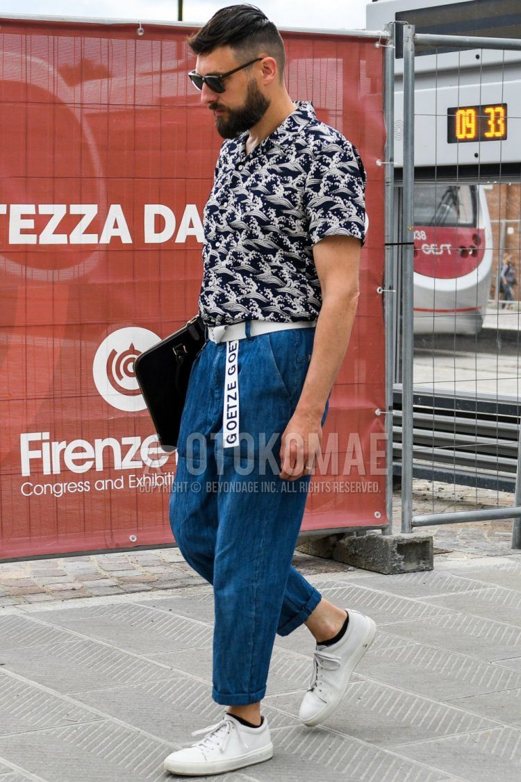 A summer men's outfit and outfit with solid color sunglasses, multi-colored graphic shirt, solid white tape belt, solid blue denim/jeans, solid blue ankle pants, white low-cut sneakers, solid black clutch/second bag/drawstring.