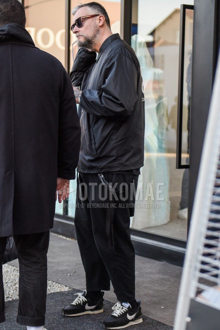 Men's fall/winter outfit with brown tortoiseshell sunglasses, solid black coach jacket, solid black slacks, solid black ankle pants, solid black socks, and Nike Undercover Daybreak black low-cut sneakers.