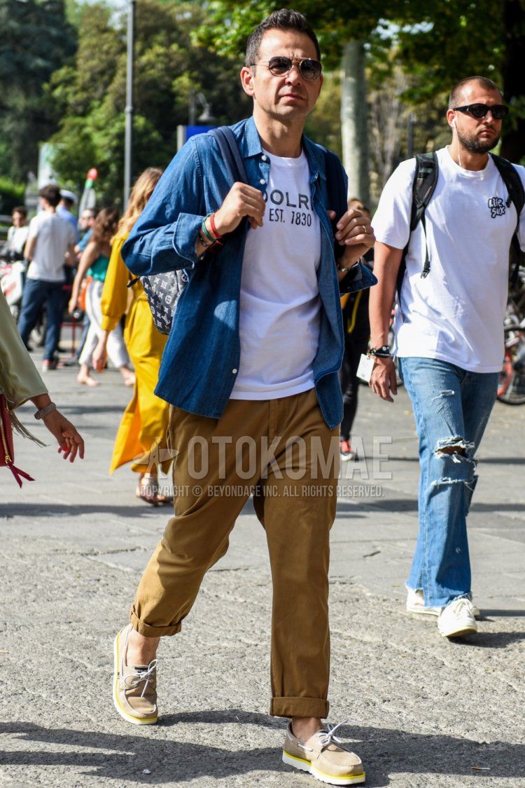 Men's spring/summer/autumn coordination and outfit with teardrop gold/black solid sunglasses, solid blue denim/chambray shirt, white top/inner t-shirt, solid brown cotton pants, and beige moccasins/deck shoes leather shoes.