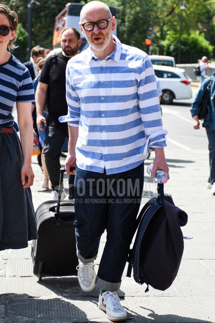 Men's spring/summer/autumn coordinate/outfit with plain black glasses, linen blue/white striped shirt, plain navy denim/jeans, white low-cut sneakers, and plain navy backpack.