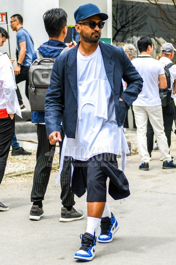 Men's spring, summer, and fall coordinate and outfit with plain gray baseball cap, plain black sunglasses, plain navy tailored jacket, plain white t-shirt, plain gray shorts, plain white socks, and Nike Air Jordan 1 white and blue high-cut sneakers.