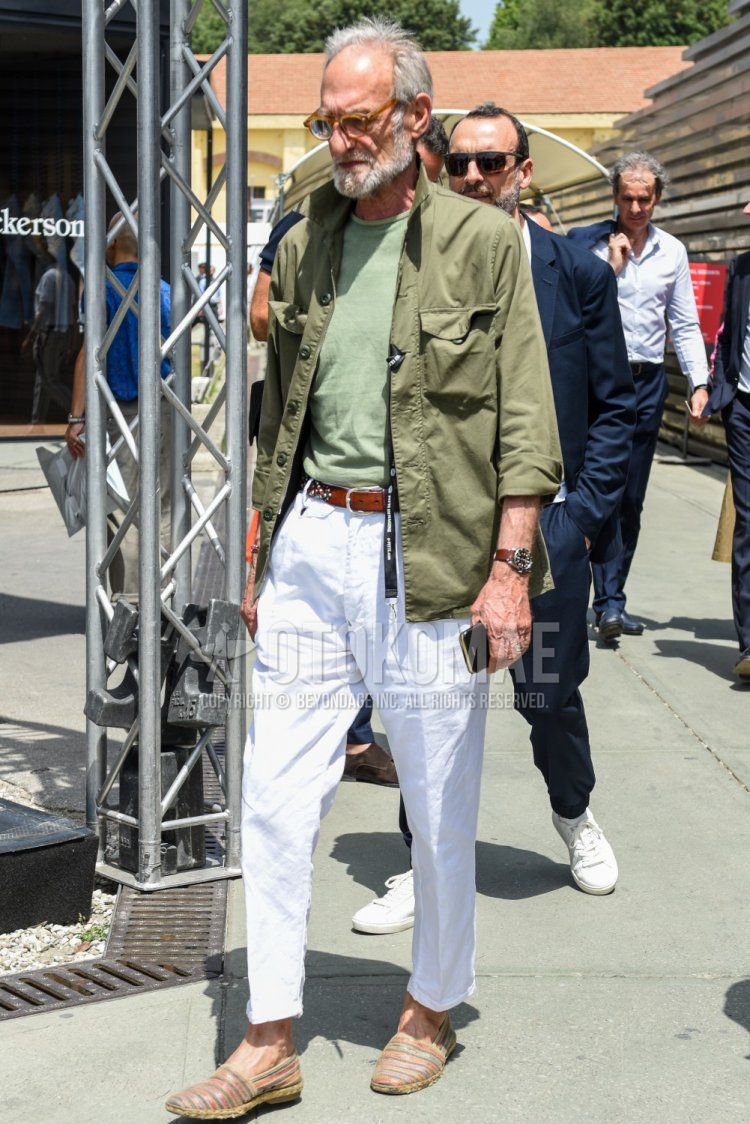 Men's spring, summer, and fall coordinate and outfit with solid beige glasses, solid olive green shirt jacket, solid olive green t-shirt, solid brown leather belt, solid white cotton pants, and multi-colored striped espadrilles.