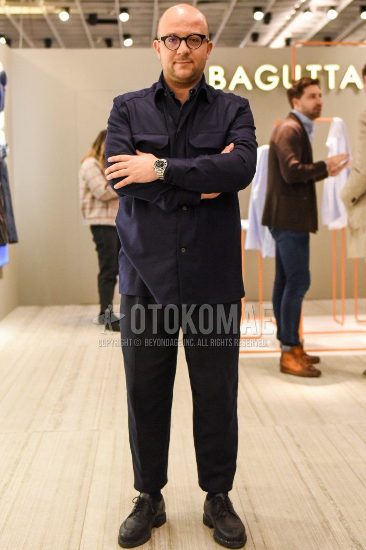 Men's spring and fall outfit with plain black glasses, plain navy shirt jacket, plain gray slacks, plain gray cropped pants, plain black socks, and brown U-tip leather shoes.