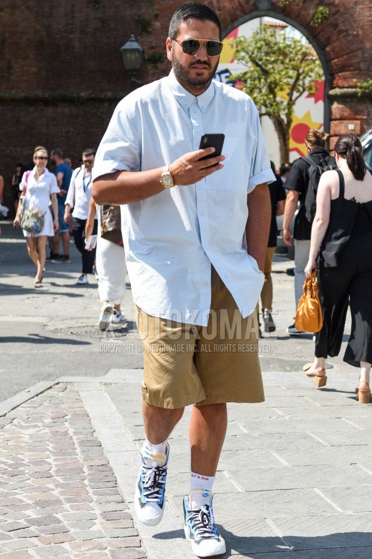 Summer men's coordinate and outfit with gold and black solid sunglasses, solid white shirt, solid beige shorts, solid white socks, and Nike Sakai light blue, white, and navy high-cut sneakers.