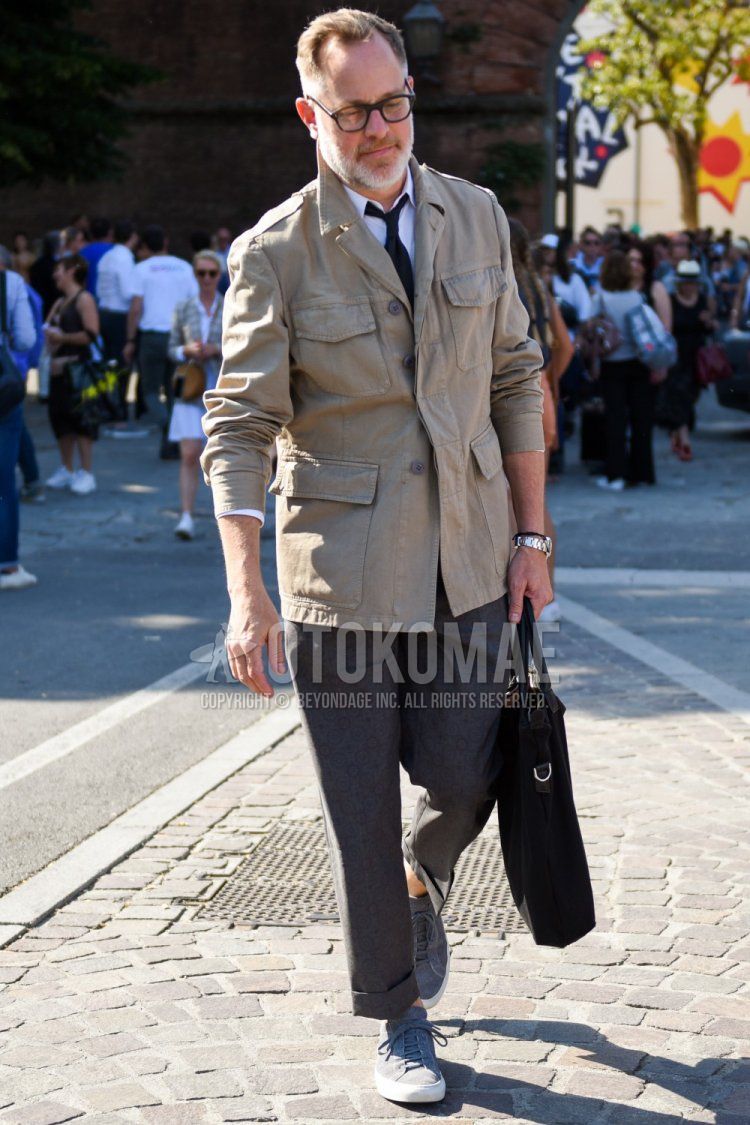 Men's spring/fall outfit with brown tortoiseshell glasses, solid beige safari jacket, solid white shirt, solid gray slacks, gray low-cut sneakers, solid black briefcase/handbag, and solid black tie.