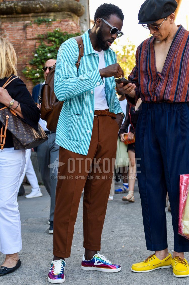 Spring and fall men's outfit with plain gray sunglasses, light blue striped seersucker shirt jacket, plain white t-shirt, plain brown mesh belt, plain leather belt, plain brown slacks, multi-colored low-cut sneakers, plain brown backpack Outfit.
