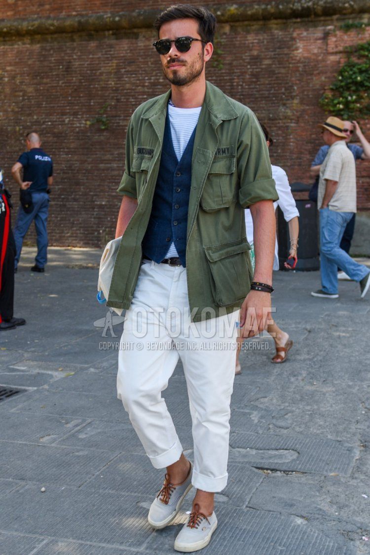 Men's spring and fall outfit with plain black sunglasses, plain olive green safari jacket, plain navy gilet, white striped t-shirt, plain white cotton pants, and white low-cut sneakers.