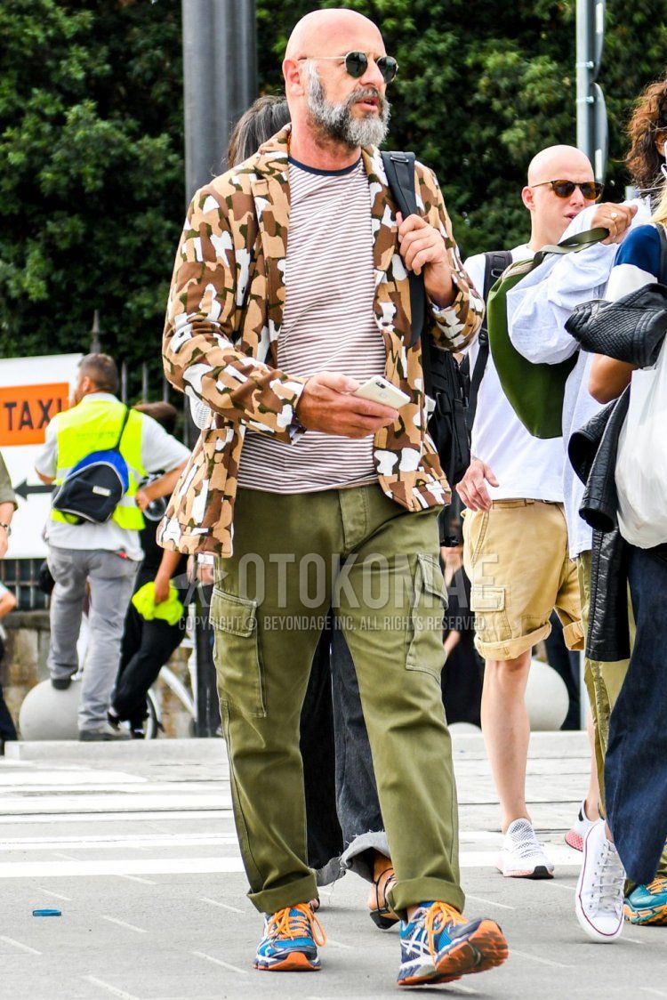 Men's spring, summer, and fall coordinate and outfit with plain gold sunglasses, brown/beige camouflage tailored jacket, white/brown striped t-shirt, plain olive green cargo pants, Asics multi-colored low-cut sneakers, and plain black shoulder bag.