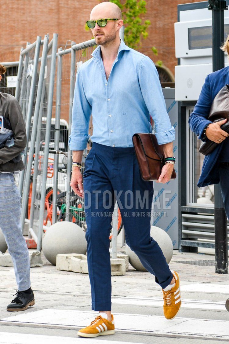 Men's spring/summer/fall outfit with solid color sunglasses, solid color light blue shirt, solid color navy slacks, Adidas yellow low-cut sneakers, solid color brown clutch/second bag/drawstring bag.
