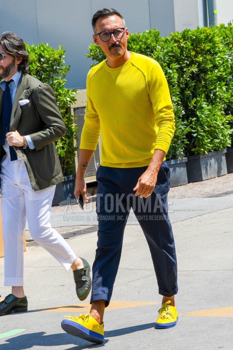 Men's spring and fall coordinate and outfit with plain glasses, plain yellow sweater, plain navy cotton pants, and yellow/navy low-cut sneakers.