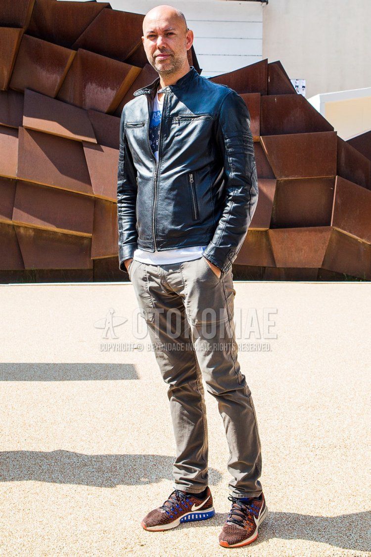 Men's spring and fall coordinate and outfit with plain black rider's jacket, white graphic t-shirt, plain beige chinos, and brown low-cut Nike sneakers.