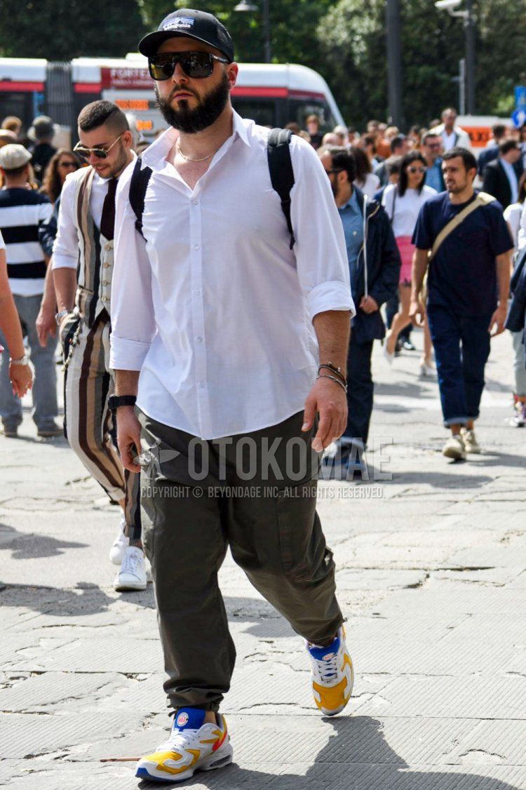 Men's spring/summer coordinate and outfit with Patagonia black decal logo baseball cap, plain black sunglasses, plain white shirt, plain olive green cargo pants, and white, yellow, and blue low-cut sneakers.