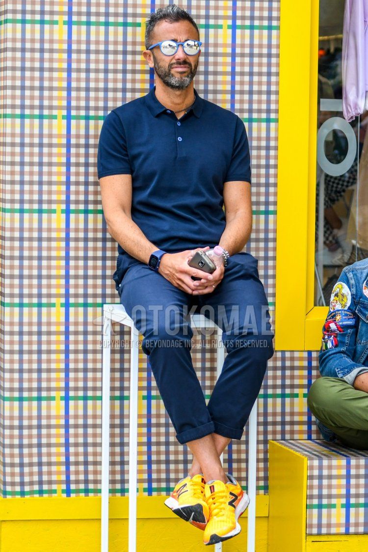 Men's spring/summer coordinate and outfit with plain blue sunglasses, plain navy polo shirt, plain navy ankle pants, and New Balance yellow/orange low-cut sneakers.