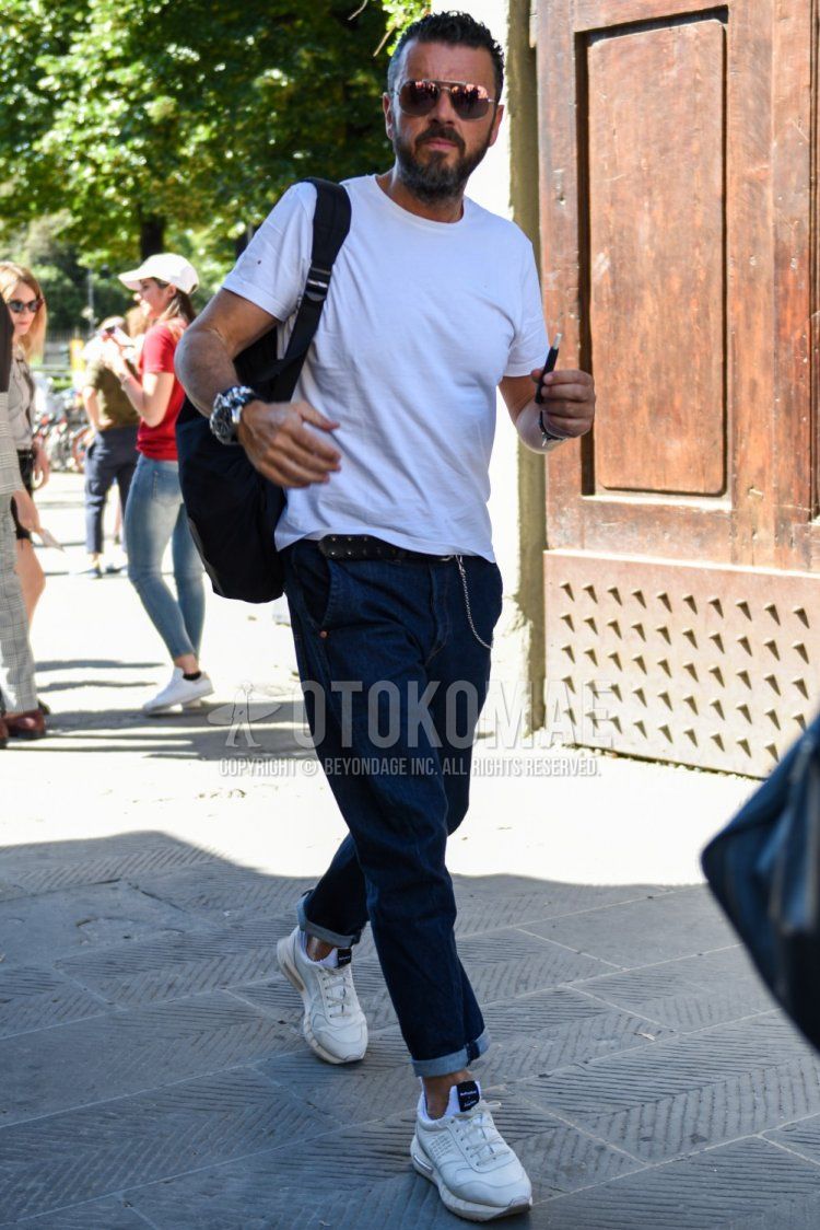 Summer men's coordinate and outfit with teardrop silver-orange solid sunglasses, solid white t-shirt, solid navy denim/jeans, white low-cut sneakers, and solid black backpack.