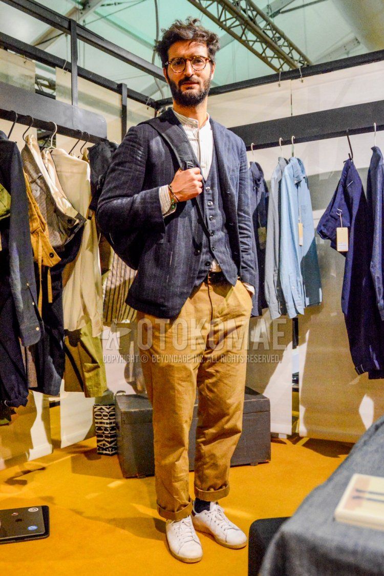 Spring and fall men's coordinate and outfit with plain glasses, navy striped tailored jacket, navy striped gilet, white striped shirt, plain black leather belt, plain beige chinos, plain black socks, and white low-cut sneakers.