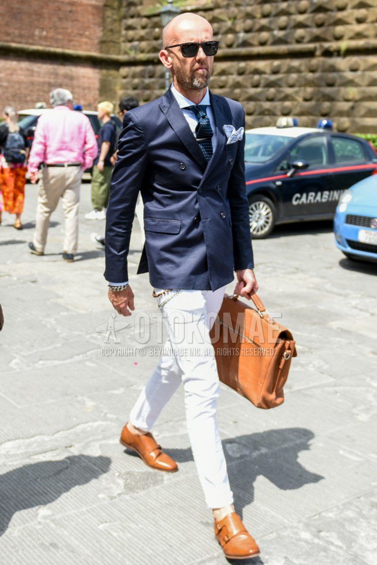 Men's spring/fall outfit with plain black sunglasses, plain navy tailored jacket, plain light blue shirt, plain white denim/jeans, brown monk shoe leather shoes, plain brown briefcase/handbag, and navy dot tie.