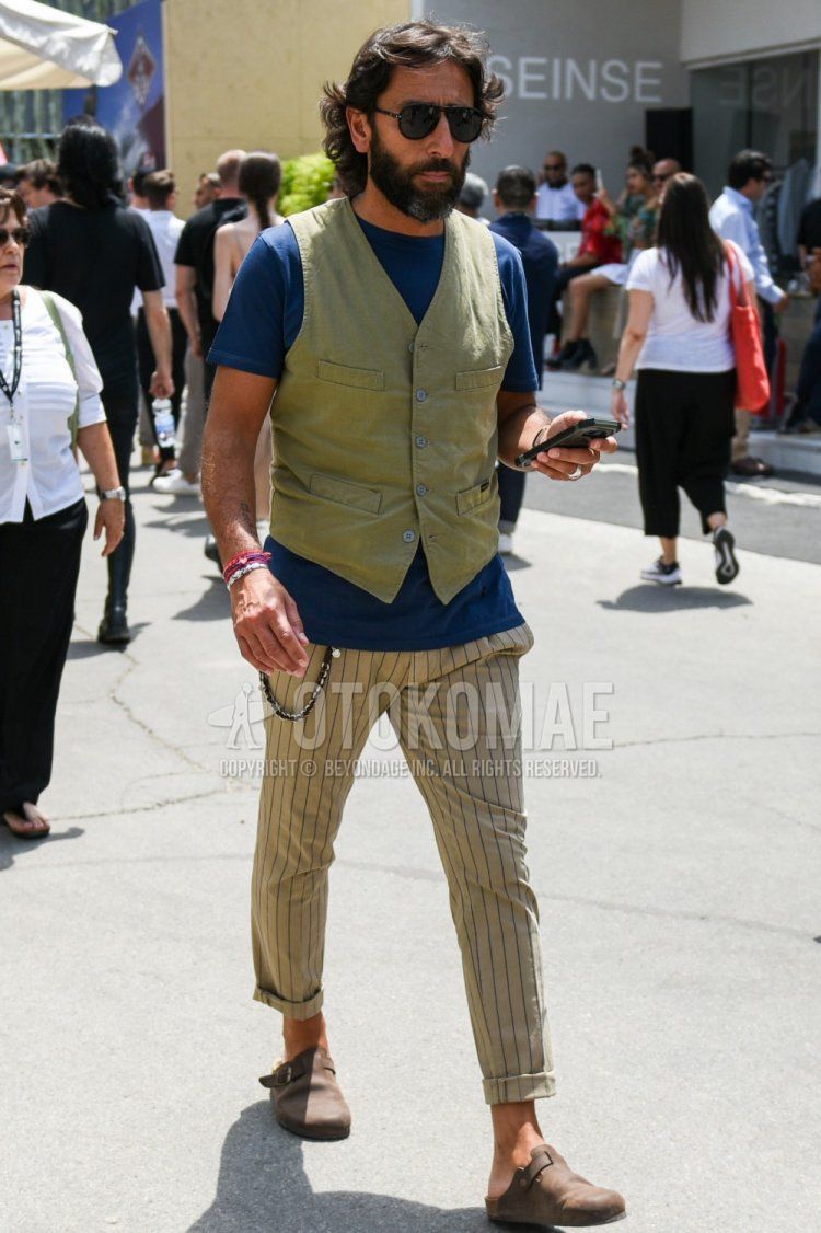 Summer men's coordinate and outfit with teardrop plain black sunglasses, olive green plain gilet, navy plain t-shirt, beige striped slacks and brown leather sandals.