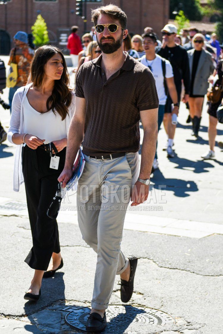 Summer men's coordinate and outfit with gold/black solid sunglasses, brown solid polo shirt, black solid leather belt, beige solid chinos and brown moccasin/deck shoes leather shoes.
