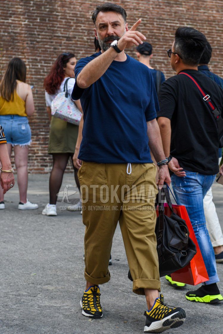 A summer men's summer outfit/coordination of a plain navy t-shirt, plain beige chinos, Nike yellow/black low-cut sneakers, and a plain black briefcase/handbag.