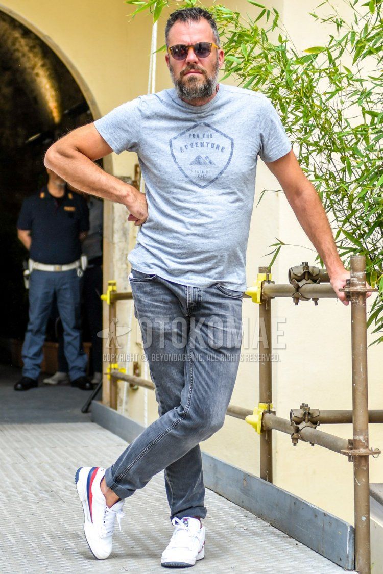 A summer men's coordinate and outfit with plain sunglasses, gray graphic t-shirt, plain gray denim/jeans, and white low-cut sneakers from Fila.